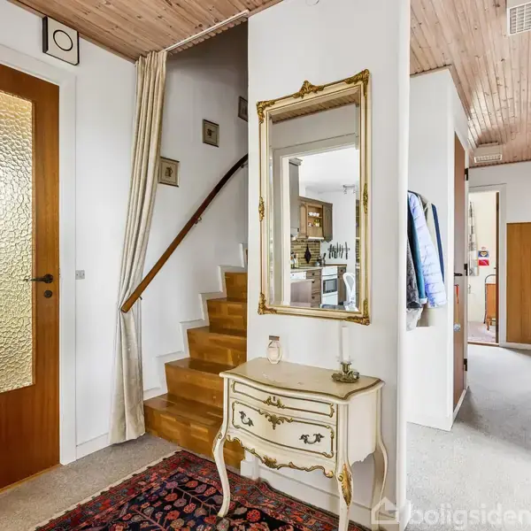 Spejl hænger over en antik kommode i en entré. Trappen fører opad ved siden af en trædør med mønstret glas. Tæppe dækker gulvet, og flere hvide vægge omfavner rummet.