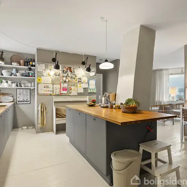 Et moderne køkken med grå skabe og trækøkkenbord, omgivet af åbne hylder med bøger og køkkengrej. På væggen hænger plakater og noter. I baggrunden ses en oplyst stue.