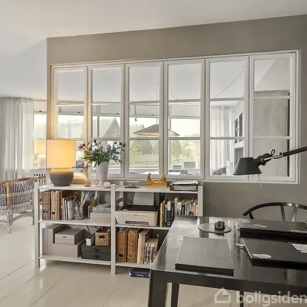 Bord med skrivebordslampe og kontorartikler står i et lyst rum med en reol fyldt med bøger. Bagerst er store vinduer med udsigt til et fællesområde.