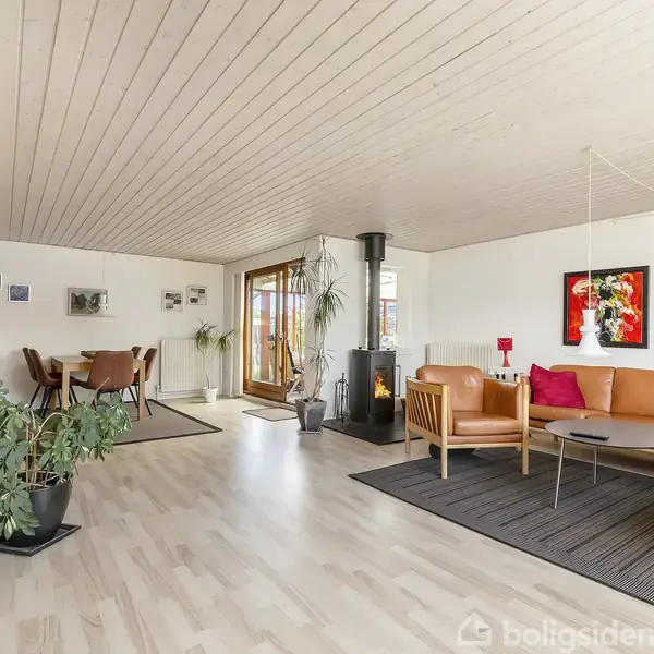 Stue med brun lædersofa og sofabord foran pejs i moderne indretning. Spisebord med stole i hjørnet, omgivet af lyse trægulve, planter og farverige malerier på væggene.
