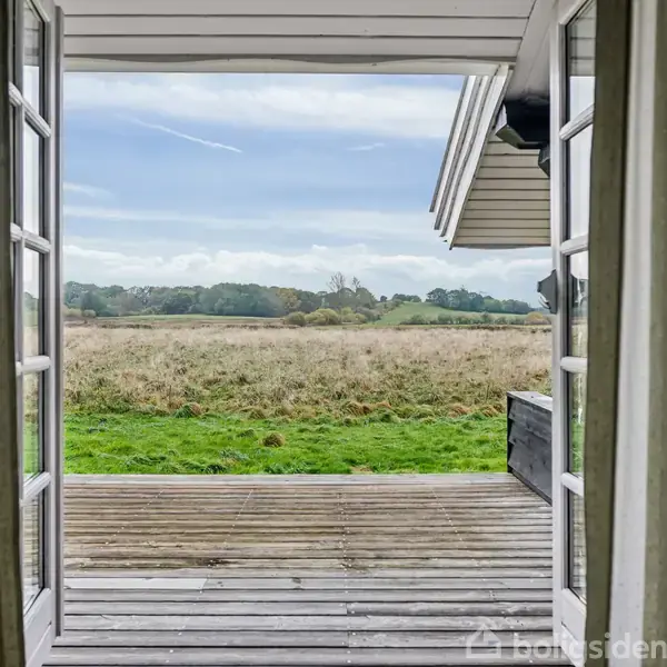 Et åbent sæt terrassedøre fører ud til en træterrasse, udsigt til en græseng og skov i horisonten, under en klar himmel. Gardiner hænger på dørens sider.