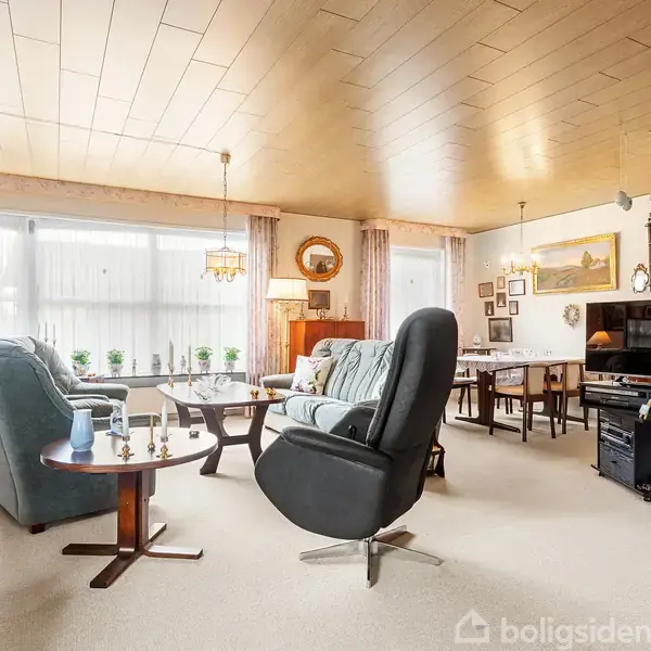 En stue med komfortable lænestole og sofabord, afslappet atmosfære med lyse gardiner og træloft. I baggrunden ses et spisebord, TV og dekorationer på væggene.