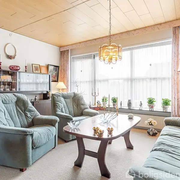 Stue med grøn sofa og to lænestole arrangeret omkring en rektangulær træ-sofabord. Store vinduer bagved med persienner; skænk med portrætter og små planter. Lysekrone hænger fra loftet.