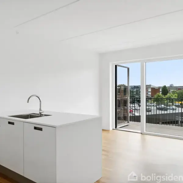 Hvidt køkkenbord med vask står i et lyst rum med trægulv. En åben dør fører ud til en balkon med udsigt til et boligområde og træer.