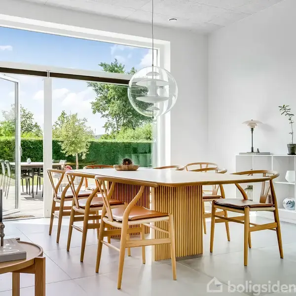 Spisebord i træ omgivet af seks stole står i et lyst rum. En åben skydedør fører ud til en terrasse med grønne buske og træer i baggrunden. Rund lampe hænger over bordet.
