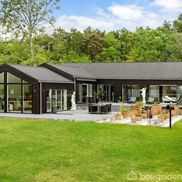Et moderne sort hus med store vinduespartier står på en flisebelagt terrasse med havefigurer og planter i potter. Omkranset af frodig græsplæne og skov, under en blå himmel med skyer.