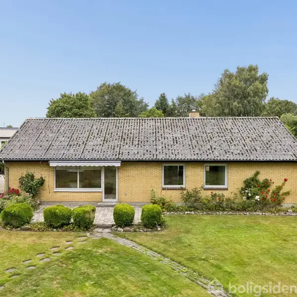 En gulstensbungalow med et gråt tegltag står midt i en velplejet have. En grussti fører op til hoveddøren, omgivet af buske og træer i baggrunden.