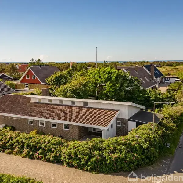 En hvid, moderne bygning med fladt tag er omgivet af grønne buske. Bygningen ligger i et villakvarter med en vej til højre og flere huse i baggrunden under en blå himmel.
