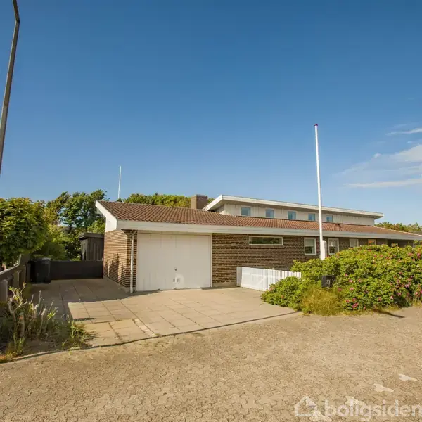 En murstensbygning med garageport står stille i en åben villavej med klart blå himmel. Grønne buske og træer omgiver huset, der har et enkelt, moderne design.
