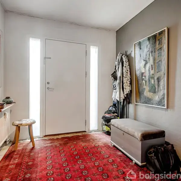 En lys entré med en rød orientalsk tæppe på gulvet. En bænk med opbevaring står under et stort billede. Frakkeophæng fyldt med tøj ved siden af en stor spejl.