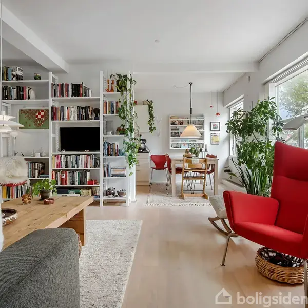 Rød lænestol står stille i lys stue med træmøbler, hylder fyldt med bøger og planter. Spisebord med lamper i baggrunden ved store vinduer.