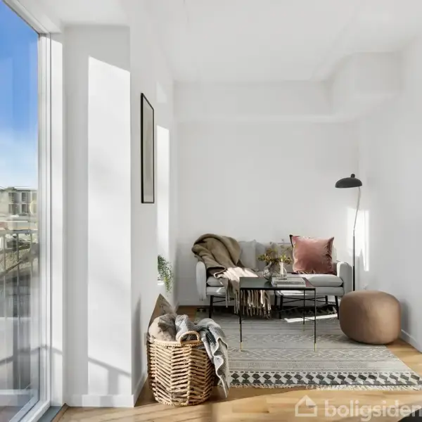 En stue med en grå sofa, en plaid og pude. Ved siden af er en gulvlampe og et sofabord. Store vinduer til venstre lader naturligt lys ind i rummet.