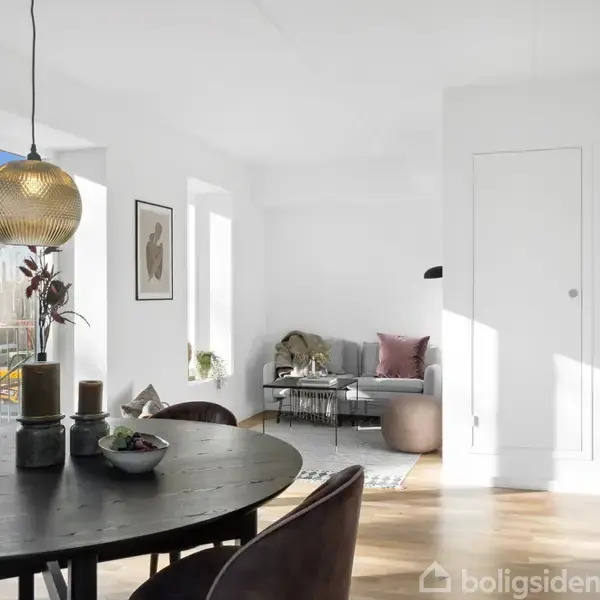 Spisebord med dekorationer står i en moderne, lys stue. I baggrunden ses en grå sofa med puder og en enkel plante foran et stort vindue.