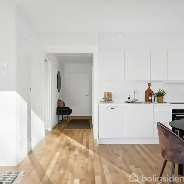 Et moderne køkken med hvide skabe og træbordplade. En sort spisebord med matchende stole står i nærheden. Rummet er lyst med trægulv og minimal dekor i en åben planløsning.