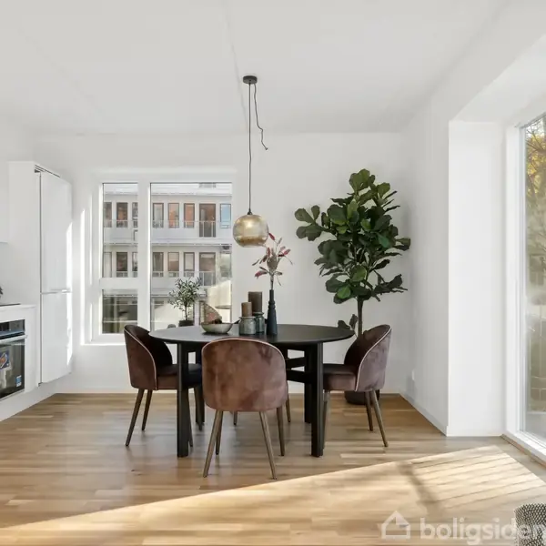En moderne spisestue med et rundt bord og fire stole. Bordet er dekoreret med blomster og et lysophæng. Rummet har trægulv, store vinduer og en plante i hjørnet.