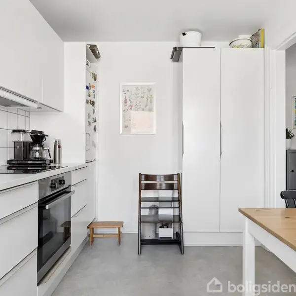 Et moderne køkken med hvide skabe og apparater. En kaffemaskine står på bordpladen. Et spisebord ses mod højre. Væggene er hvide, og gulvet er gråt. En stol står ved væggen.