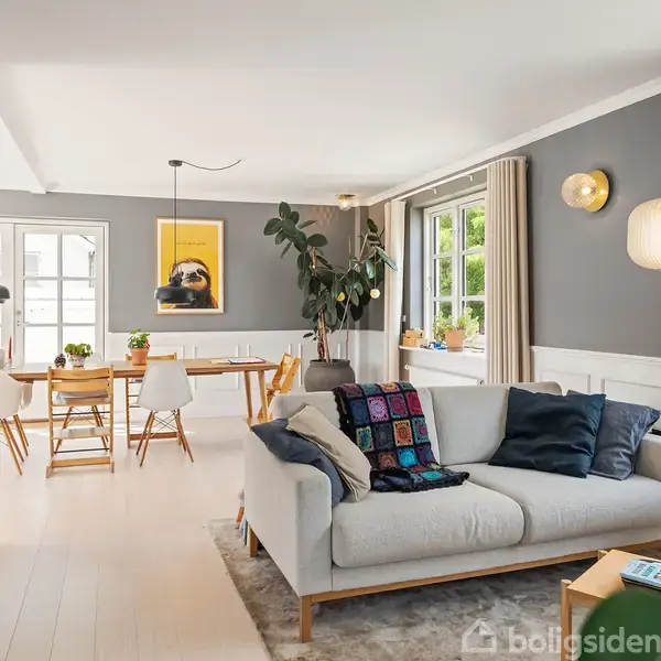En stue med en lys sofa dekoreret med farverige tæpper står på et tæppe. I baggrunden ses et spisebord med træstole, og store vinduer giver naturligt lys.