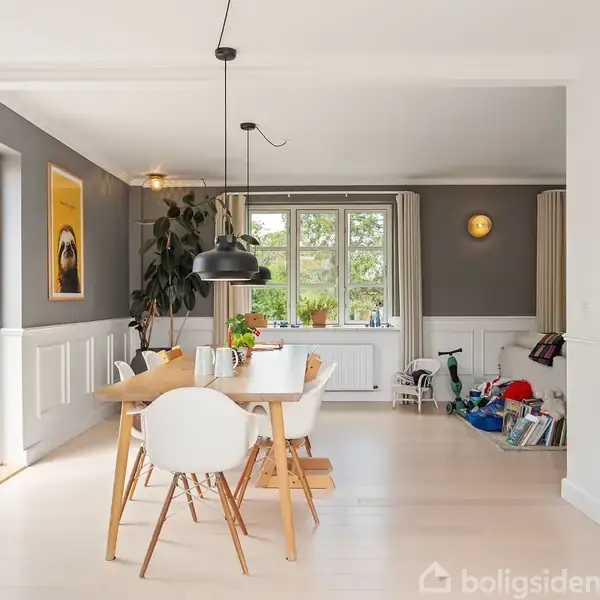 Spisebord står i et lyst, moderne rum med hvide stole. Over bordet hænger en sort lampe. Til venstre ses en åben dør, til højre legetøj og en reol. Tegning på væggen med teksten "Jun".