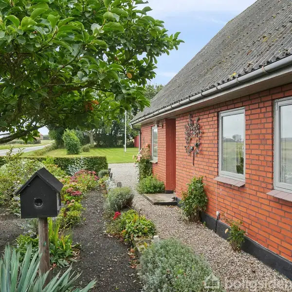Et rødt murstenshus står i en velplejet have med buske og blomster. Fuglekasse hænger på en stolpe. Sti fører til en dør. Området er landligt.