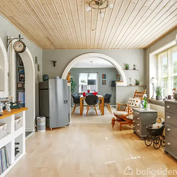 Et lyst rum med trægulv og træloft. En buegang fører til et spiseområde med bord og stole. Rummet indeholder også kommode, planter og en gyngestol ved vinduet.