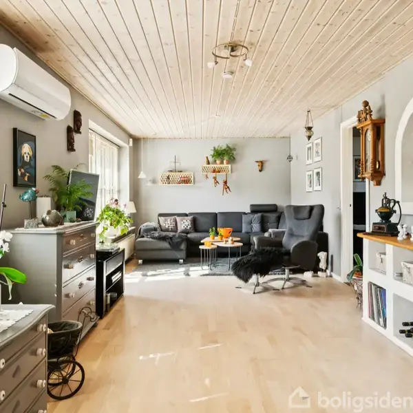 Stue med sofaarrangement og bord, omgivet af grønne planter og indretningsgenstande på træhylder. Køligt lys trænger ind gennem store vinduer, der oplyser rummet. Kunst og ure pryder væggene.
