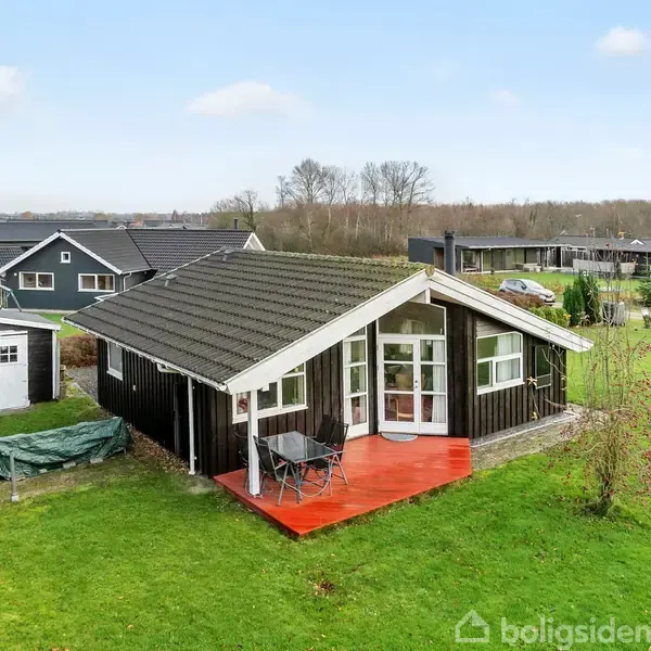 Et lille sort træhus står på en grøn græsplæne med en rød veranda foran. Ved siden af er et hvidt skur. Baggrunden viser flere huse og træer.