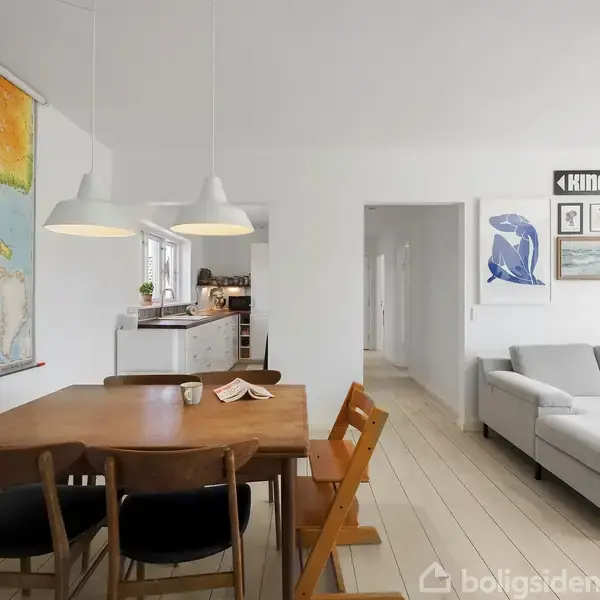 Træbord omgivet af stole og hængende lamper, placeret nær et vægkort. Stuen har hvidmalede vægge med kunstværker. I baggrunden ses en lys grå sofa og køkkenadgang.