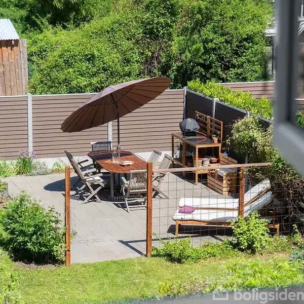 En terrasse med træbord og stole under en stor parasol. Ved siden af er der en liggestol og en grillplads. Omkring er der en have med græs og buske.