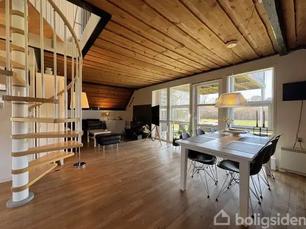 En lys stue med træbeklædt loft og gulv, rummer en hvid spiraltrappe, spisebord med stole, sofaområde med lænestole og lamper, store vinduer giver udsigt til udendørsområde.