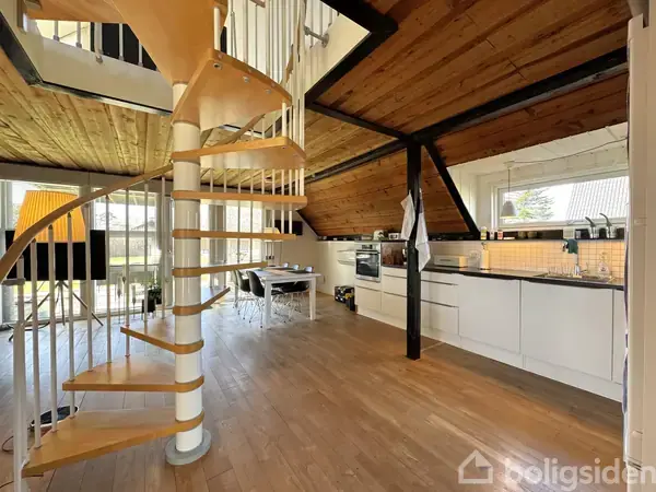 Vindeltrappe i træ snor sig opad i et moderne køkken med skrå lofter. Køkkenet har hvide skabe, mørk bordplade og et stort vindue med udsigt til træer.