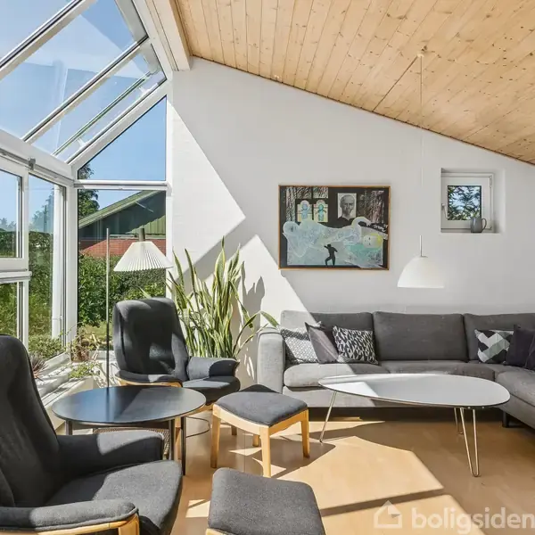 Stue med sorte møbler, sofa og lænestole omkring et hvidt bord. Store vinduer og ovenlys slipper sollys ind, omgivet af planter og vægkunst. Lyse trælofter giver en varm atmosfære.