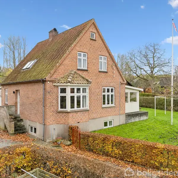 Et rødt murstenshus med en trekantet taget, omgivet af en velplejet græsplæne og hæk. I baggrunden ses træer, en flagstang samt en blå himmel med få skyer.