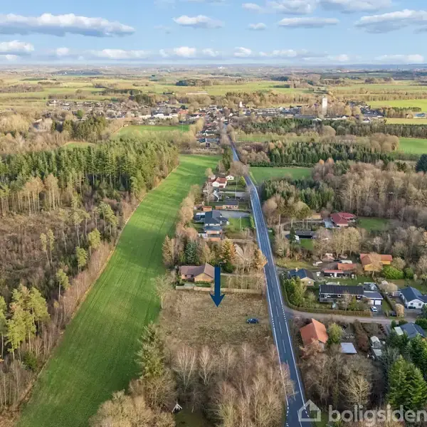 En pil peger mod en åben, græsbevokset mark omgivet af spredte huse og skov. Det omkringliggende landskab består af marker og træer under en blå himmel med få skyer.
