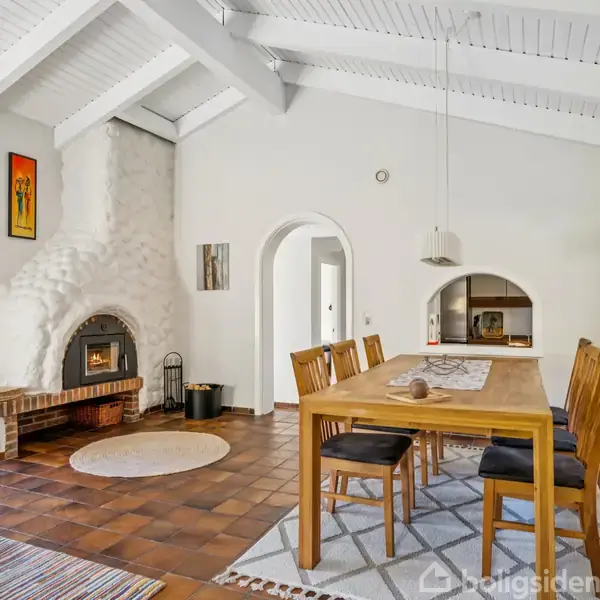 En stor spisebord med træstole står i et lyst rum med hvidmalede bjælker og en buet, dekorativ pejs. Væggene er pyntet med farverige malerier, og store vinduer lukker lys ind.