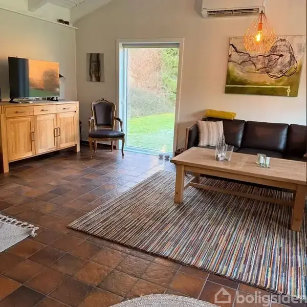 En stue med en lædersofa og et sofabord på et stribet tæppe. Til venstre står et fjernsyn på et træmøbel. En stor glasdør fører til en have.