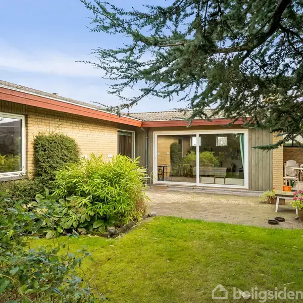 Et etplanshus med store vinduer og en terrasse. Der er en terrasse med havemøbler i en have med tætte buske og en velholdt græsplæne.