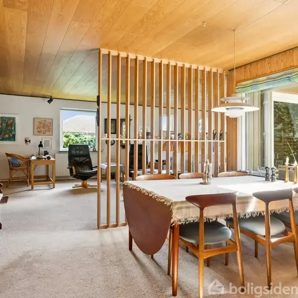 En spisebord med seks stole står i en stue med træloft og skillevæg i trælameller. Et klaver står til venstre, og store vinduer giver udsigt til haven.