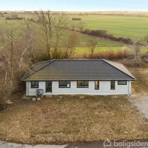 Hus står isoleret på en grusgrund, omgivet af marker. Det har et mørkt tag og lyse ydervægge, med nogle få træer i nærheden og en nabo på højre side.
