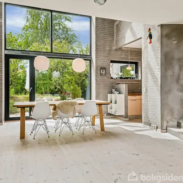 En lys, moderne spisestue med et stort træbord omgivet af seks hvide stole. Store vinduer skaber en åben atmosfære med udsigt til en grøn have. Ingen tekst fundet.