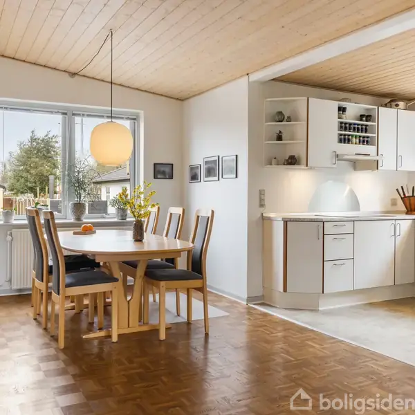 Spisebord med seks stole står under en rund lampe i en spisestue. Der er en plante og kunstværker på væggen. Køkken med hvide skabe ses i baggrunden.