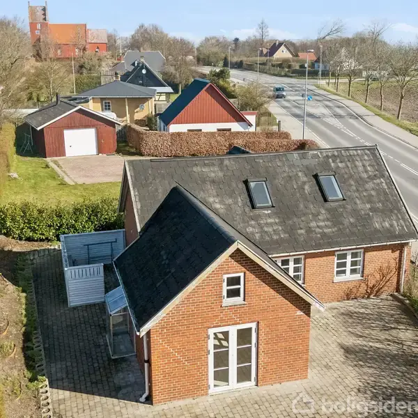 Rødt murstenshus med skråt tag og tagvinduer står stille ved en brolagt gårdsplads. Omkringliggende kontekst inkluderer en landevej og flere nabobygninger samt en hæk som skiller ejendommen.