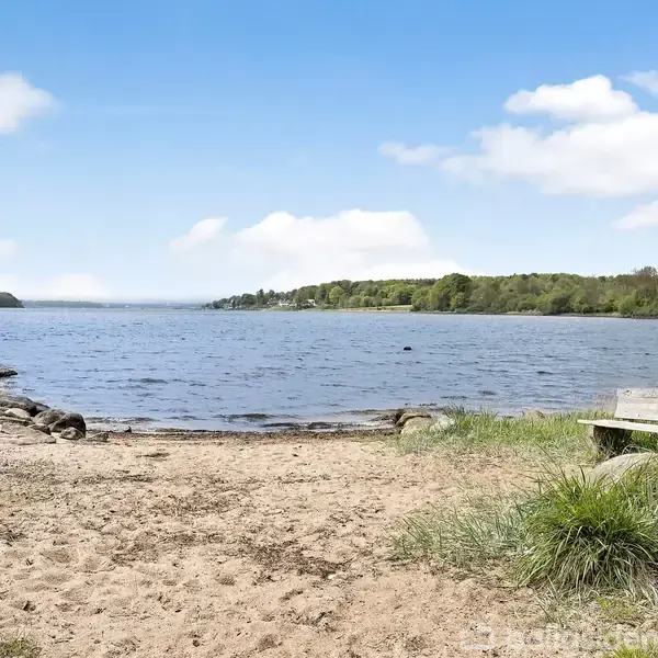 En sandstrand skuer ud over en stille sø med en træbænk placeret på græsset til højre. Skov og kystlinje er synlige i baggrunden under en blå himmel med skyer.