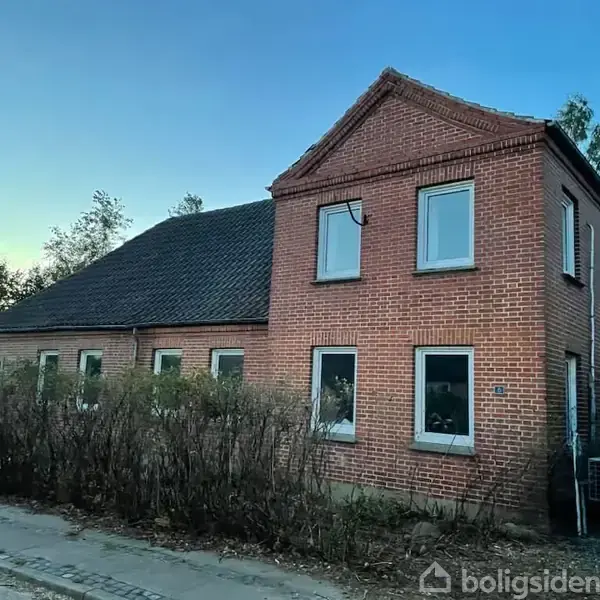 Murstenshus med fem vinduer står stille, omgivet af lav buskbevoksning og træer i en villavejssammenhæng med en blå himmel i baggrunden.