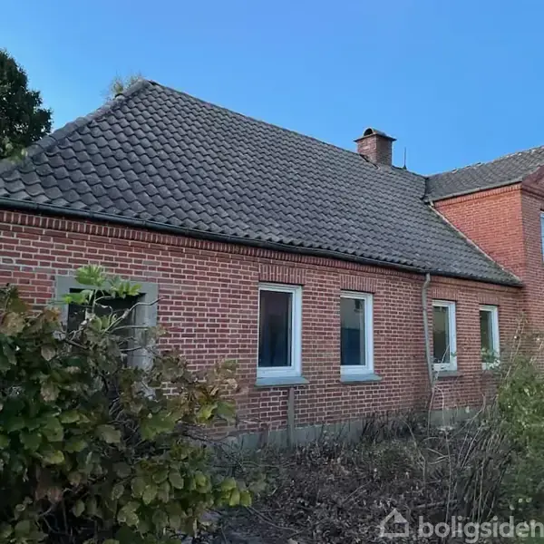 Rødt murstenshus med skråt tag, flere vinduer og en skorsten. Omkring huset er der tæt bevoksning med buske og træer under en klar himmel.