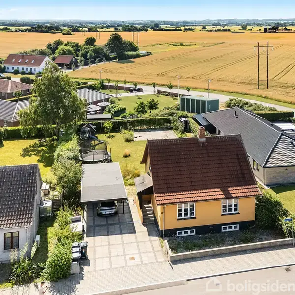 Et gult hus står i et kvarter ved siden af andre huse. Der er en indkørsel og have. Baggrundslandskabet består af marker og grønne områder under en klar himmel.