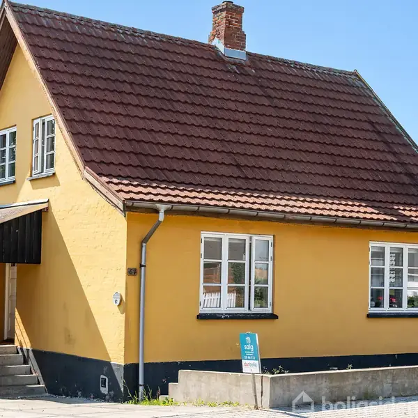 Et gult hus med rødt tegltag står i en solrig gårdsplads. Der er en skorsten og flere vinduer. Et skilt foran huset viser teksten "Til salg 139 Bolig".