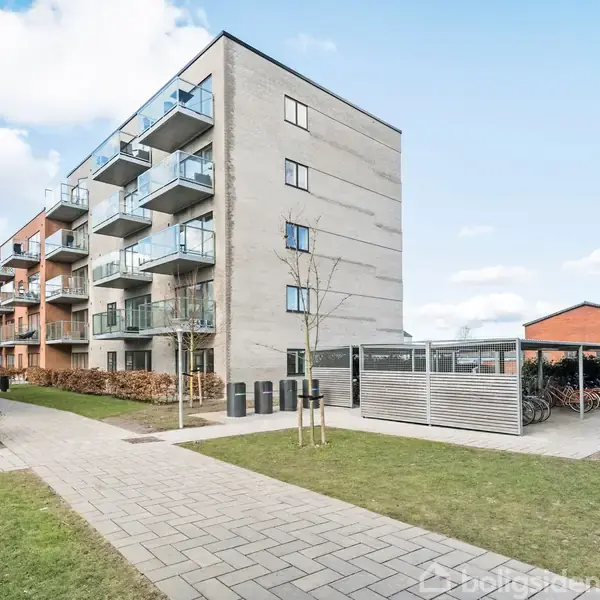 En moderne, firkantet lejlighedsbygning med balkoner står på en flisebelagt sti med græsplæner omkring. I forgrunden ses en cykelparkering med overdækning og skraldespande.
