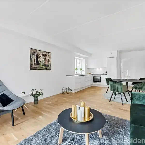 En moderne stue med en grå lænestol, et rundt sofabord med dekorationer, og en grøn sofa i et lyst rum. I baggrunden ses et åbent køkken og spiseområde.