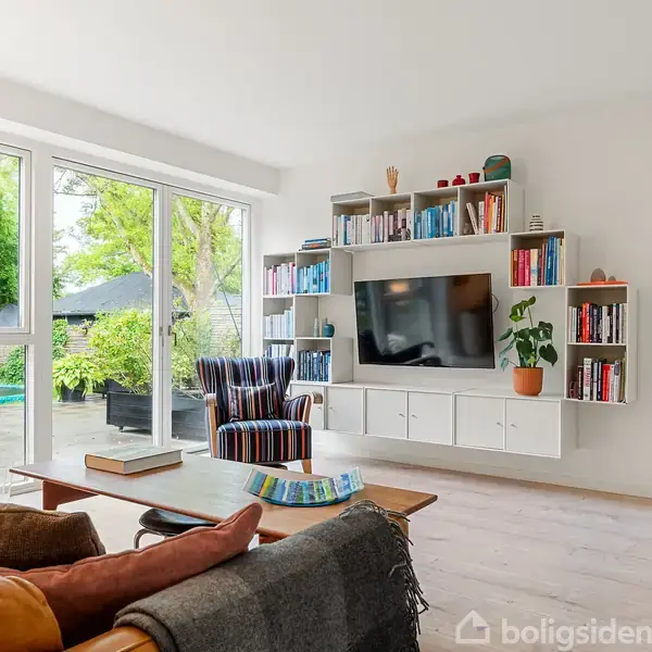 En stue med sofa og stol omkranser et sofabord; væghængt tv med reol omkring. Store vinduer viser en terrasse med planter og træer udenfor.