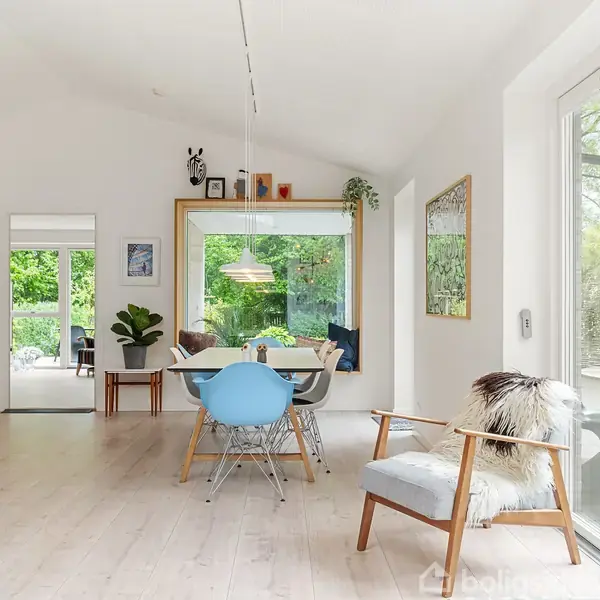 Stue med spisebord midt i rummet, omgivet af stole. Store vinduer og glasdøre åbner mod frodig have. En lampe hænger over bordet, og der er moderne indretning.